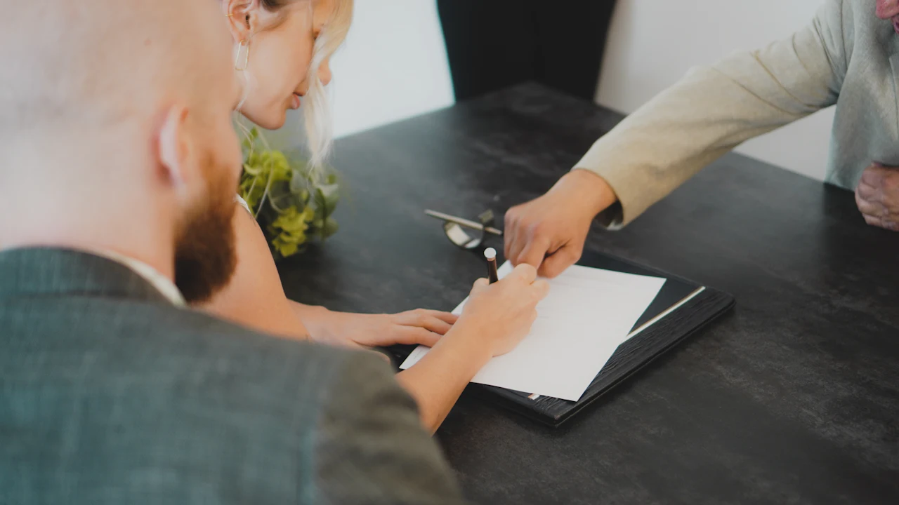 Two people leaning over a dark desk to sign a document while a third person points at the page