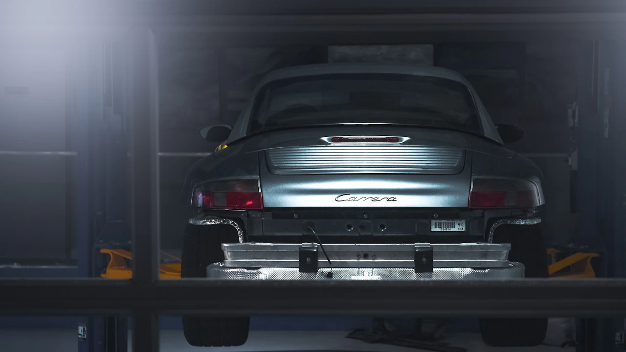 A silver sports car raised on a workshop lift, photographed from behind with its underside and exhausts visible