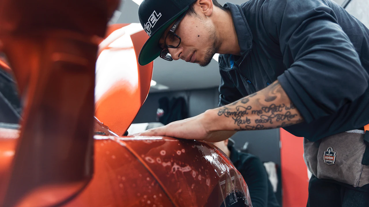 A person in a cap leaning close over the wing of an orange car, examining the paint surface at a shallow angle