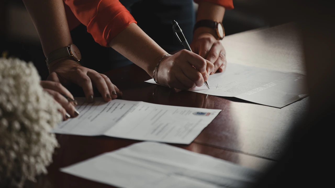 Two pairs of hands over printed forms on a polished dark table, one person signing while the other rests a hand on a second document