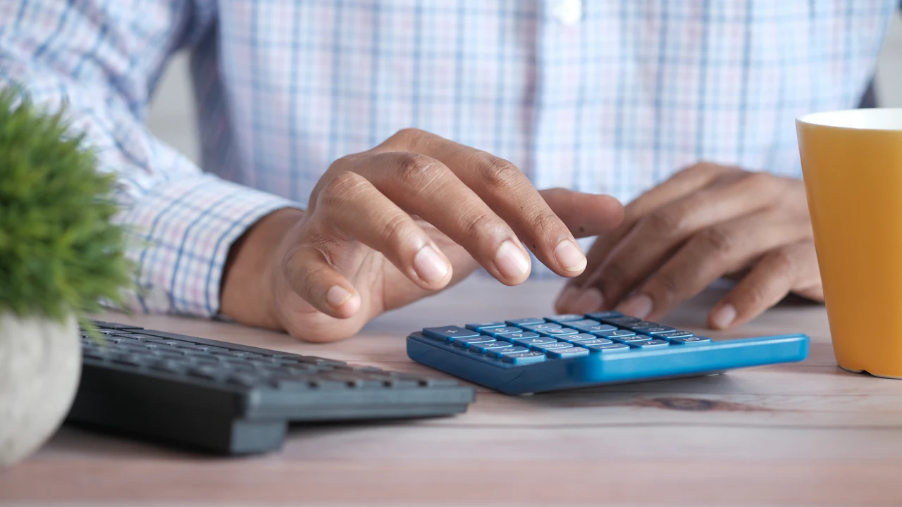 A man in a checked shirt working a blue desktop calculator, a keyboard and a yellow mug on the desk beside him