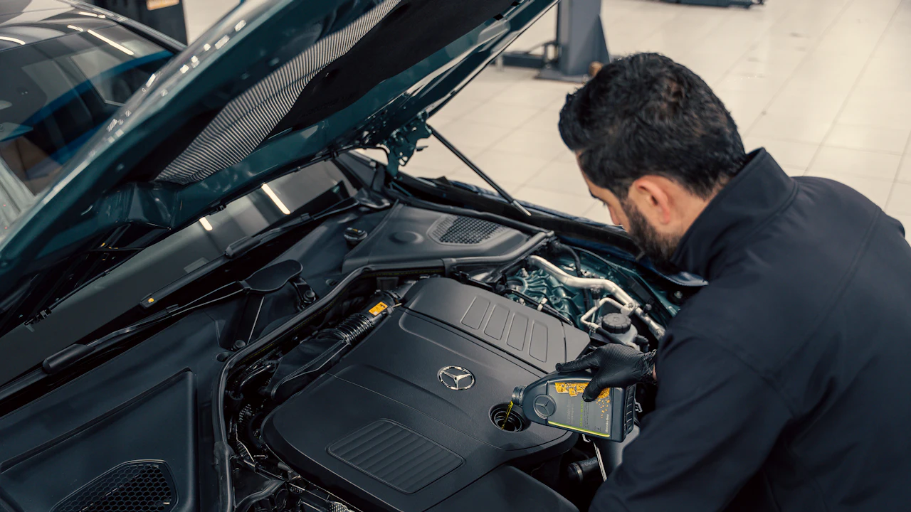 A person leaning under the raised bonnet of a dark saloon in a workshop, working on the engine bay