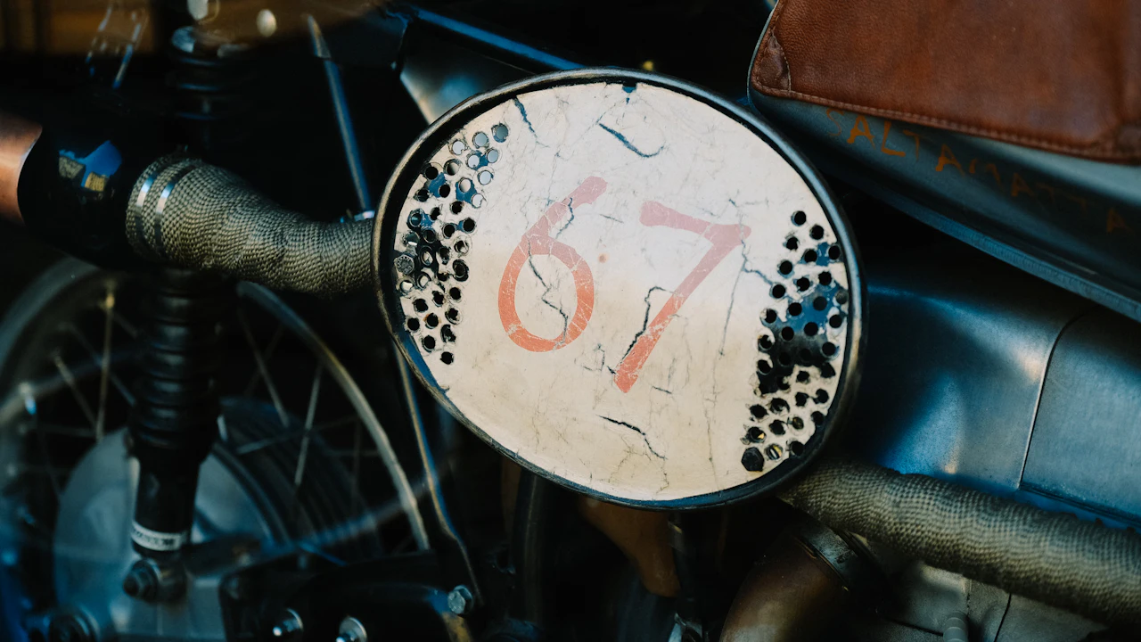 A vintage racing motorcycle photographed close up, a cracked and weathered oval number board mounted beside the forks above a spoked front wheel