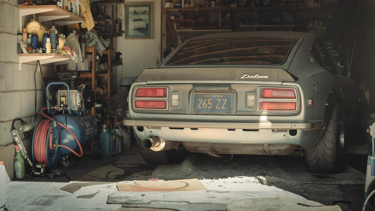 A dusty coupé parked in a home garage beside an air compressor, hose reel and shelves of workshop supplies