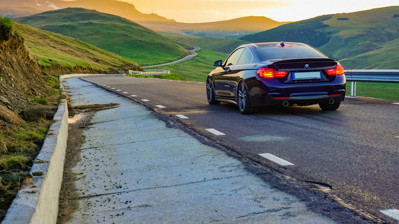 A dark coupe seen from behind on an empty mountain road at sunset, the tarmac curving away between green hillsides