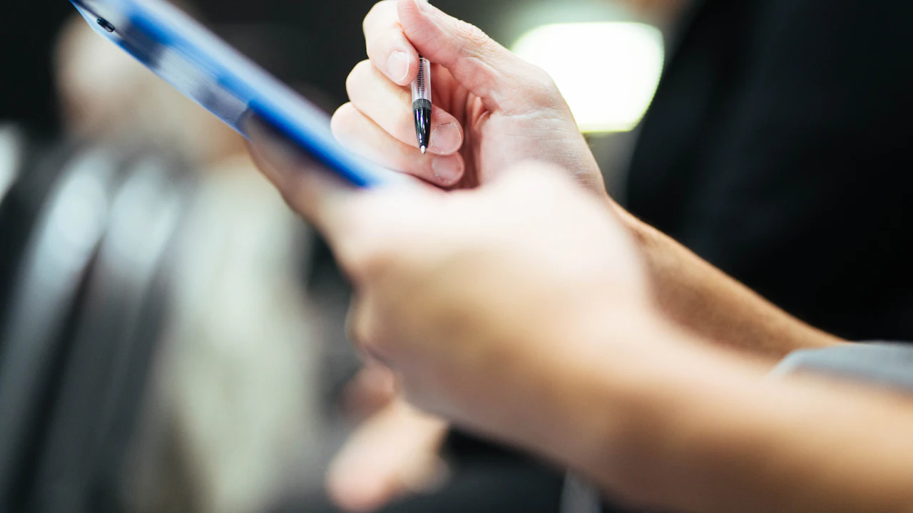 A close-up of a hand holding a ballpoint pen against a clipboard, the paperwork out of focus in the foreground under warm indoor light