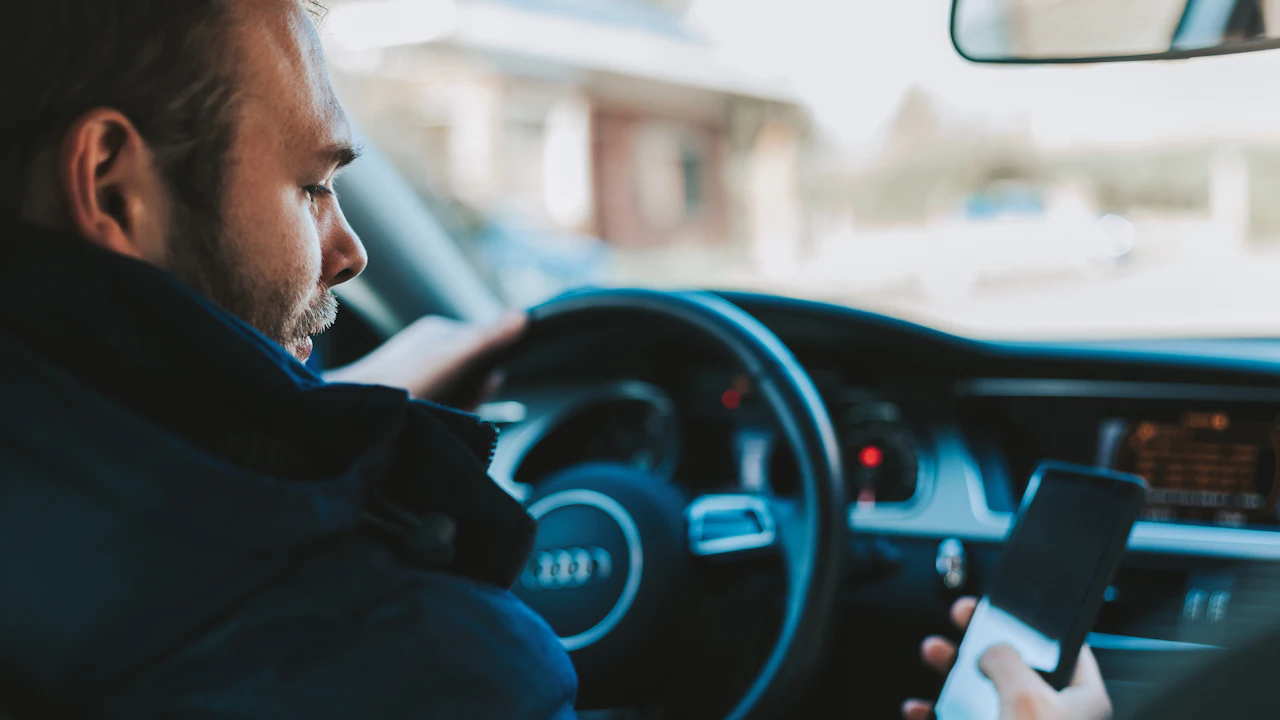 A man sitting in a car’s driver seat holding a phone in one hand, the steering wheel and lit instrument cluster in front of him