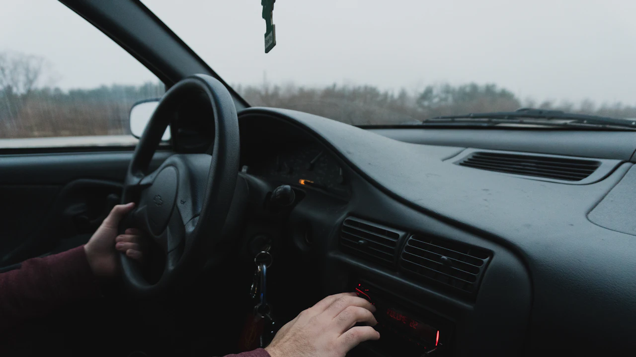 The interior of an older car seen from the passenger seat, the driver’s hands on the wheel and the stereo, keys hanging from the ignition barrel