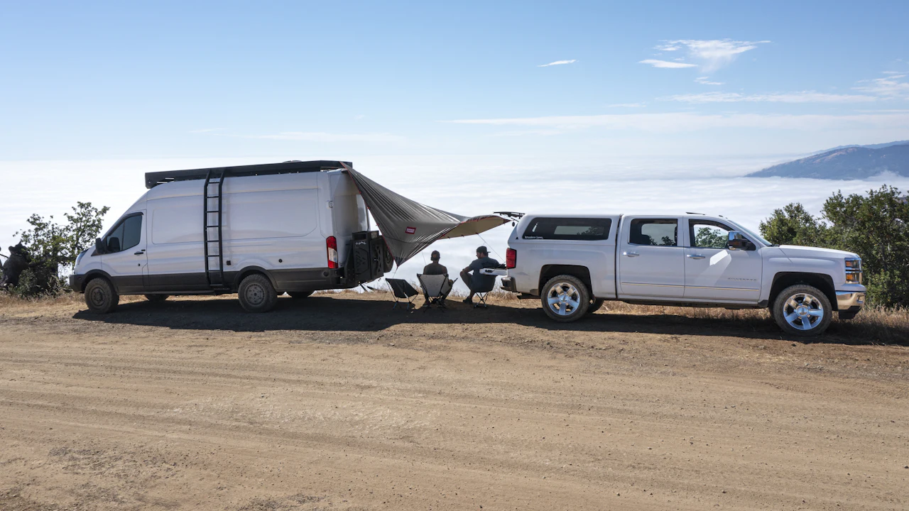 A white pickup truck and a van with a side awning parked on a dirt clearing above a bank of low cloud, with people sitting between them
