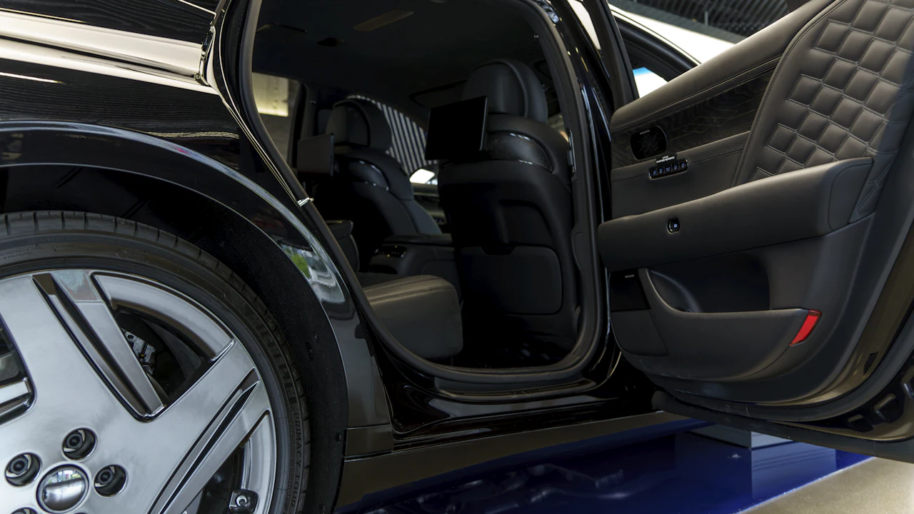 The rear door of a black luxury saloon standing open on a showroom floor, showing quilted black leather seats and the door jamb, with a polished multi-spoke alloy wheel in the foreground