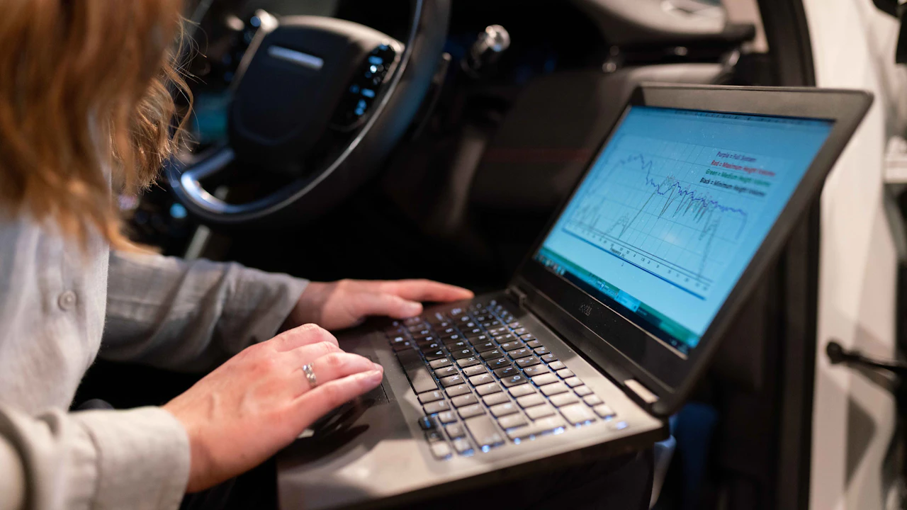 A technician working at a laptop propped on a stand inside a car, a diagnostic trace running across the screen beside the steering wheel